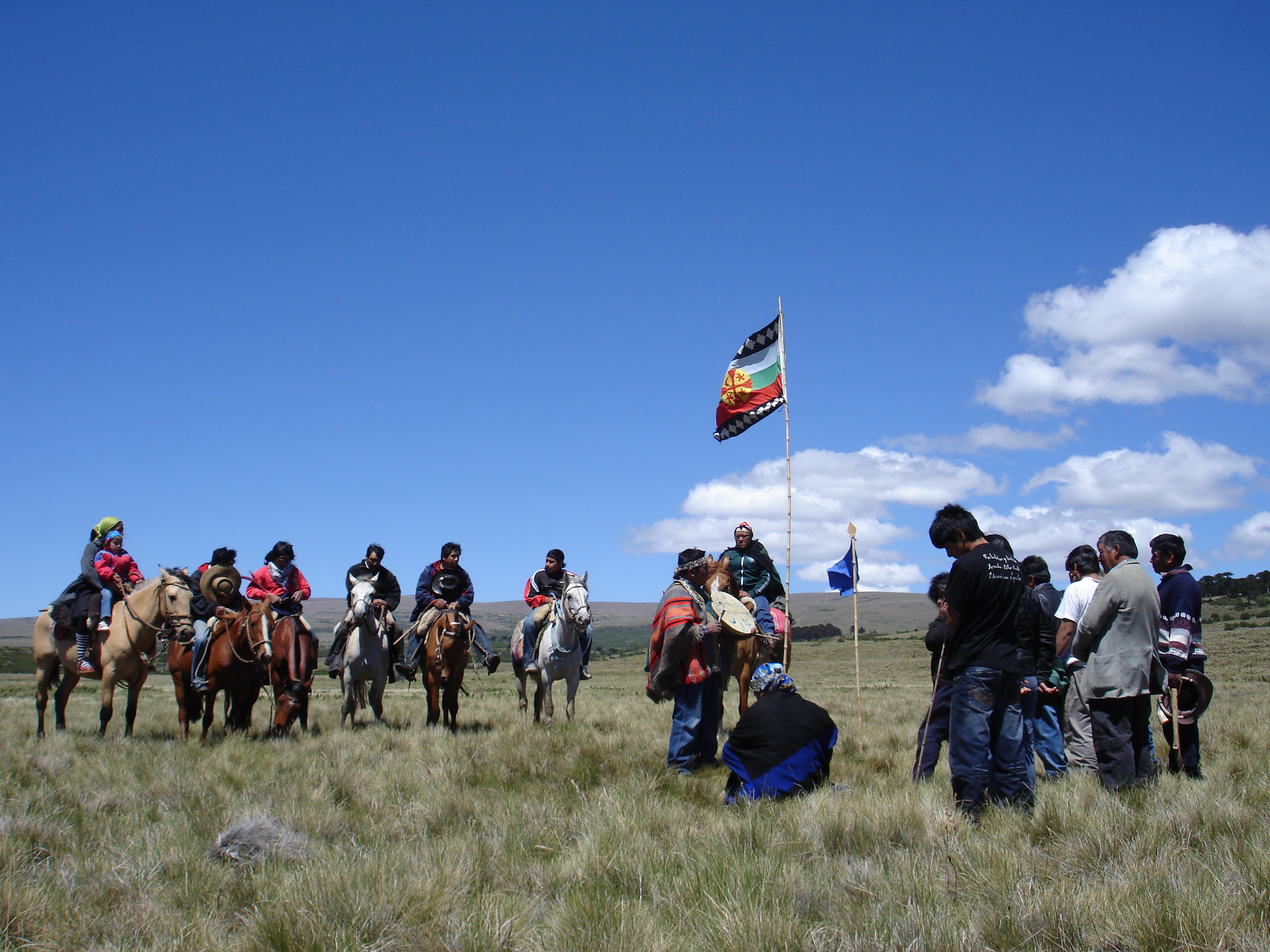 The Mapuche flag flies over a group of people, some on horseback and others standing or sitting in a circle, during a religious ceremony held in a grassy area under a blue sky