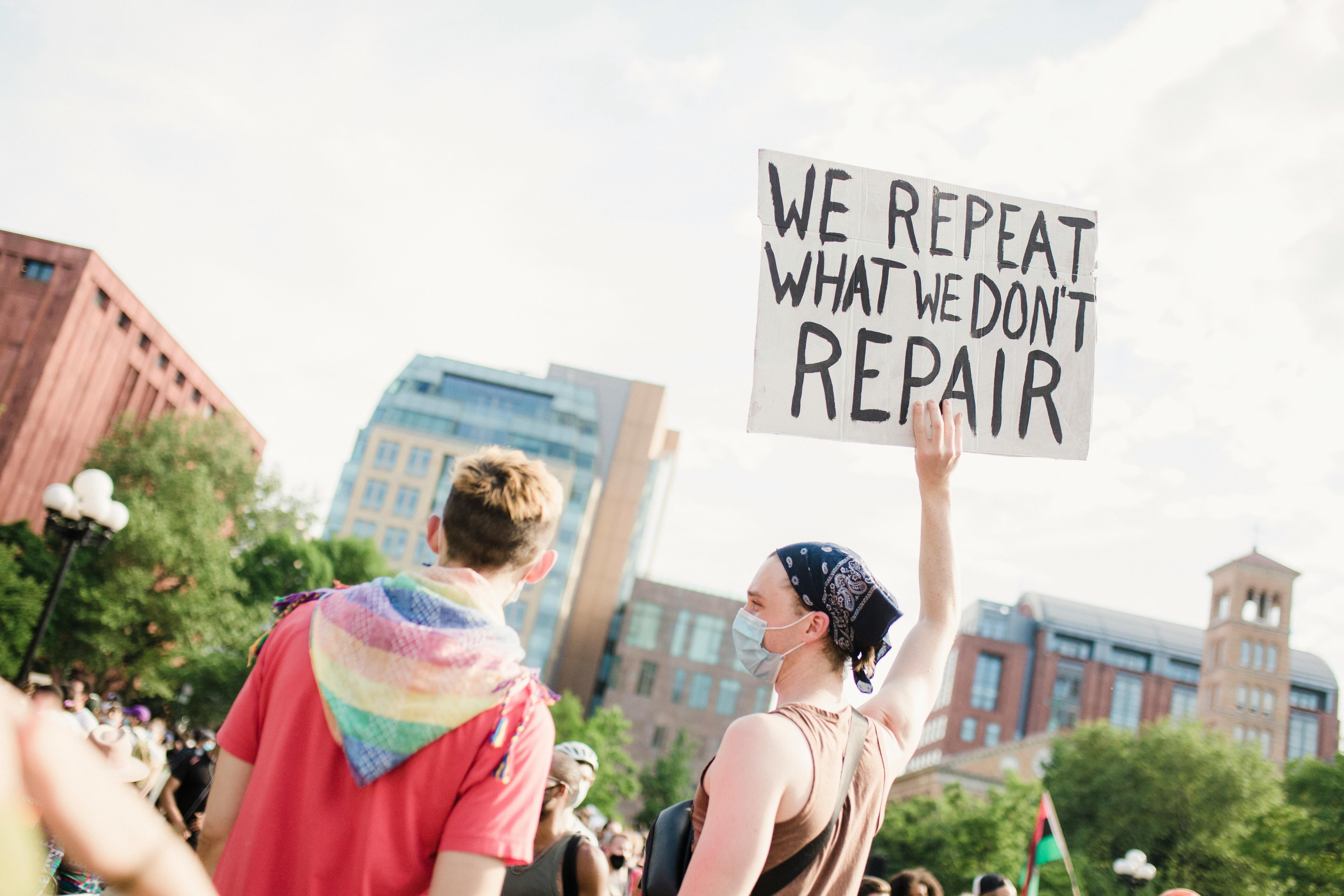 Two people at a protest facing away from the camera. One is holding a sign that reads "we repeat what we don't repair"